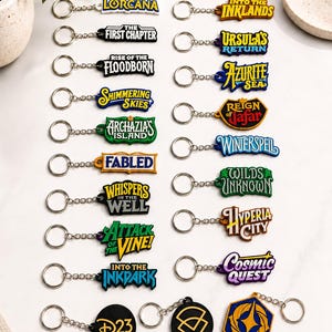 May include: A collection of twenty-one colorful rubber keychains featuring various logos and text including Lorcana, Into the Inklands, The First Chapter, Ursula's Return, Rise of the Floodborn, Azurite Sea, Shimmering Skies, Reign of Jafar, and D23.