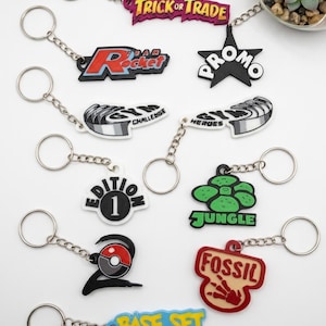 Może przedstawiać: Kolekcja dziesięciu gumowych breloków z różnymi logotypami i napisami, w tym Trick or Trade, Team Rocket, Promo, Gym Challenge, Gym Heroes, Edition 1, Jungle, ikoną Poké Ball, Fossil oraz Base Set, każdy przymocowany do srebrnego metalowego kółka.