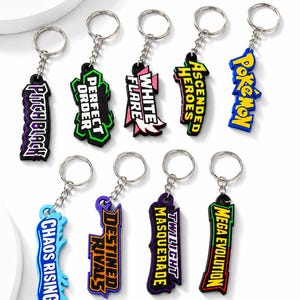 May include: A collection of nine colorful rubber keychains featuring various text designs including Pitch Black, Perfect Order, White Flare, Ascended Heroes, Pokémon, Chaos Rising, Destined Rivals, Twilight Masquerade, and Mega Evolution with metal rings.