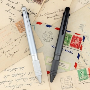 May include: Two multi-function pens, one in a matte silver finish and the other in a matte charcoal gray finish. Each pen features a textured grip section near the tip, a pocket clip, and a top-mounted mechanism for selecting different ink colors.