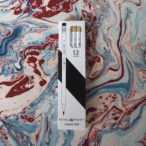 Puede incluir: Una caja blanca de lápices Royal Point Janus 929, con un diseño diagonal en blanco y negro. La caja contiene 12 lápices HB/#2. Los lápices se muestran sobre un fondo veteado con remolinos rojos, azules y blancos.