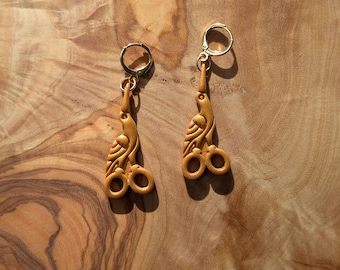 May include: A pair of gold-colored earrings with a bird and scissors design. The earrings are made of a light brown material and have a lever back closure. The earrings are hanging on a wooden surface.