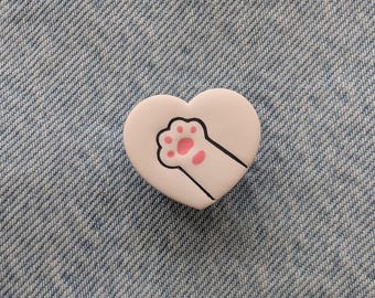 May include: A white heart-shaped pin with a black outline, featuring a cat paw print with pink pads. The pin is set against a blue denim background. This item is perfect for cat lovers.
