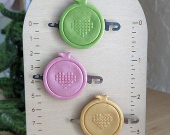 May include: A wooden stand with three embroidery hoops, each with a different color: green, pink, and gold. The hoops are attached to the stand with metal clips. The stand has a ruler on the side with numbers 0 through 9.