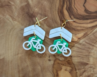 May include: A pair of green and white polymer clay earrings featuring a bicycle design. The earrings are shaped like a hexagon with a white bicycle silhouette on a green background. The earrings are attached to a white chevron-shaped piece.