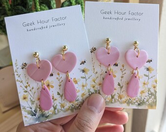 May include: Pink heart and teardrop shaped polymer clay earrings with gold hooks. The earrings are displayed on a white card with the text "Geek Hour Factor handcrafted jewellery".