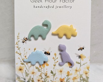 May include: Four colorful dinosaur-shaped earrings on a white card with the text "Geek Hour Factor handcrafted jewellery".
