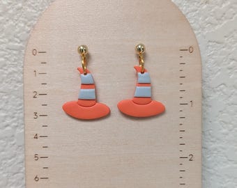 May include: A pair of dangle earrings shaped like witch hats, featuring orange and light blue horizontal stripes. The earrings are attached to gold-tone metal posts and are displayed on a wooden card with a ruler showing a length of 1.5 inches.