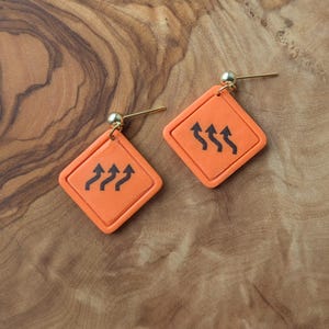 May include: A pair of orange diamond-shaped earrings with a black arrow design. The earrings have a gold-colored post and a small gold ball at the top. The earrings are displayed on a wooden surface.