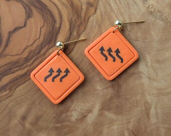 May include: A pair of orange diamond-shaped earrings with a black arrow design. The earrings have a gold-colored post and a small gold ball at the top. The earrings are displayed on a wooden surface.
