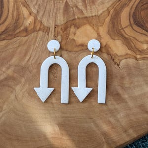 Puede incluir: Un par de pendientes de cerámica blanca en forma de U invertida con una flecha apuntando hacia abajo. Los pendientes tienen una textura moteada y ganchos dorados.