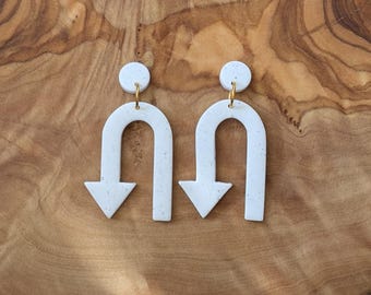 May include: A pair of white ceramic earrings shaped like an upside down U with an arrow pointing down at the bottom. The earrings have a speckled texture and gold hooks.