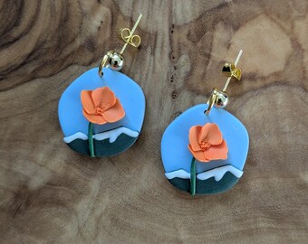 May include: A pair of handmade earrings featuring a light blue background with an orange flower and green stem. The earrings have a gold-colored post and a small gold ball. The design includes white accents resembling snow.