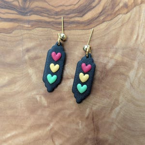 Puede incluir: Un par de pendientes negros colgantes con tres dijes en forma de corazón rojo, amarillo y verde.