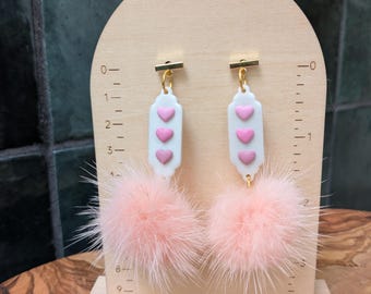 May include: A pair of pink and white earrings with three pink hearts on each earring. The earrings are attached to a pink fluffy pom-pom.