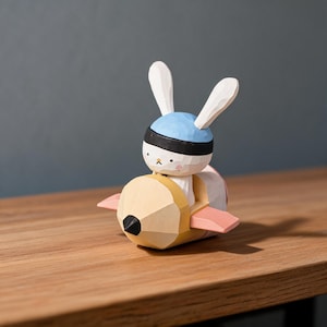 May include: A carved wooden figurine of a white rabbit with long ears, wearing a blue cap and black headband, seated inside a yellow pencil-shaped vehicle with small pink wings on the sides and a black pointed tip at the front.