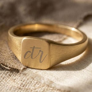 May include: A gold-colored signet ring with a square face. The face of the ring has the engraved letters "ctr" in a cursive font. The ring is made of a brushed metal and sits on a neutral-colored fabric.