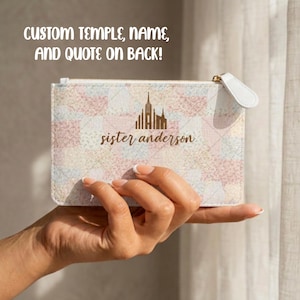 May include: A rectangular clutch bag with a gold zip and white pull tab. The front displays a temple illustration and the name "sister anderson." Text at the top reads "CUSTOM TEMPLE, NAME, AND QUOTE ON BACK!"