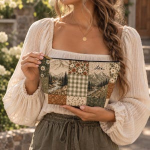 May include: A rectangular zippered pouch featuring a patchwork design of mountain landscapes, pine forests, green gingham, and floral patterns in earth tones. The fabric surface includes the embroidered name Lexi in black cursive script.