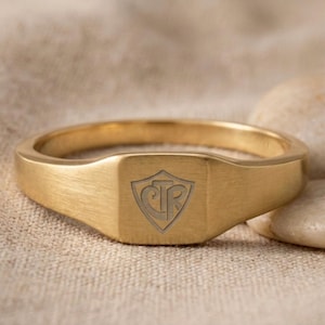 Puede incluir: Un anillo de sello dorado con acabado cepillado. El anillo presenta una cara rectangular con un diseño de cresta grabado. La cresta tiene las letras "CTR" dentro de un escudo. El anillo se muestra sobre una superficie de tono neutro.