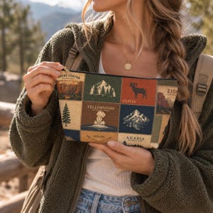 Op de afbeelding: Een rechthoekig tasje met rits voorzien van een patchworkontwerp van diverse nationale parken. De print bevat illustraties van bergen, bossen, een dikhoornschaap en geisers, met tekstlabels voor Grand Canyon, Yellowstone, Zion en Acadia.