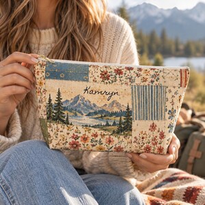 Puede incluir: Un estuche rectangular con cremallera que presenta un diseño tipo patchwork con motivos florales y una ilustración central de montañas, un lago y pinos. El nombre Kamryn está impreso en letra cursiva negra sobre el paisaje montañoso.