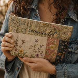 May include: A rectangular zippered pouch featuring a patchwork design of floral patterns in shades of beige, pink, yellow, and blue. The word savannah is printed in black cursive script across the upper section of the fabric pouch.