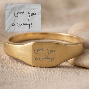 May include: A gold signet ring with the inscription "Love you always" engraved on the face. The ring is displayed on a neutral-toned surface, with a handwritten note that reads the same text in the background.