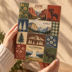 Könnte beinhalten: Ein rechteckiges Notizbuch mit festem Einband und einer Collage aus bedruckten Feldern. Das Cover zeigt Illustrationen des Zion-Nationalparks, des Yellowstone-Nationalparks, Berglandschaften, Kiefern, Wildblumen, ein Dickhornschaf und einen Hirsch. Der Name Emily ist auf dem Cover aufgedruckt.