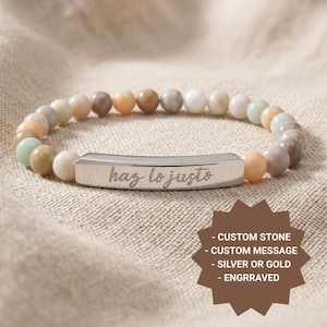 May include: Beaded bracelet with a silver bar engraved with "haz lo justo". The bracelet features a mix of neutral-toned beads in shades of beige, peach, gray, and green. The bracelet is displayed on a neutral fabric background.