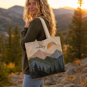 Puede incluir: Un bolso de tela color crema con un diseño de paisaje montañoso acolchado con telas superpuestas en tonos azul, verde y bronceado. La palabra oakley está bordada en cursiva negra en la parte superior izquierda del bolso, que tiene dos asas.