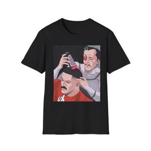 May include: Black t-shirt featuring a cartoon illustration of two men. One man is cutting the other's hair with a trimmer. The man getting his hair cut is wearing a red shirt and has a large mustache.
