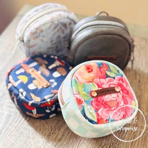 Könnte beinhalten: Vier kleine, runde Reißverschlusstaschen mit verschiedenen Mustern. Eine hat einen Blumendruck, eine andere ein Pilzdesign, eine dritte ein Blättermuster und die letzte ist ein dunkelgraues, lederartiges Material. Jede Tasche hat ein kleines Lederetikett mit "handmade".