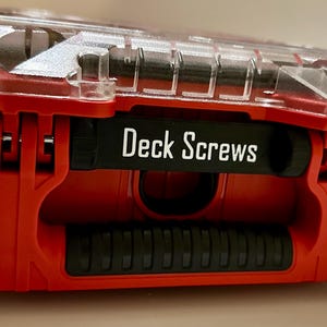 Può includere: Un contenitore rosso e nero con coperchio trasparente. Il contenitore riporta la scritta "Deck Screws" su un'etichetta nera. Il contenitore è progettato per contenere viti e altri piccoli articoli di ferramenta.