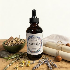 Puede incluir: Una botella de vidrio marrón con un cuentagotas negro, etiquetada como "Parasite Liquid Herbal Extract", se encuentra sobre una superficie de madera. La botella está rodeada de hierbas secas, un pequeño cuenco verde y pergamino enrollado. La etiqueta incluye el texto "Moorberry Apothecary".