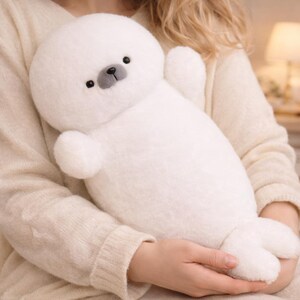 May include: A soft, white plush seal toy with a round head, small black eyes, and a gray nose. The stuffed animal has small flipper-like arms and a tail. The toy is made of a fluffy material.
