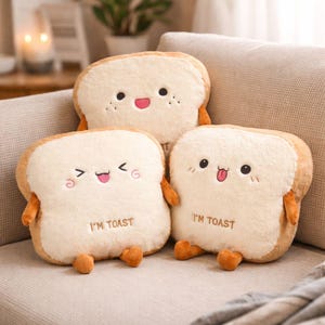 Peut inclure: Trois coussins en forme de tranche de pain avec des visages souriants et le texte "I'M TOAST". Les coussins ont une croûte beige et un centre blanc cassé doux. Ils sont disposés sur un canapé beige, créant une scène chaleureuse et ludique.