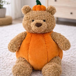 Peut inclure: Un ours en peluche vêtu d'un costume de citrouille orange avec une tige verte. L'ours a une fourrure brune et un nez brun. Le costume de citrouille recouvre le corps de l'ours et a une forme ronde. L'ours est assis sur un tapis blanc.