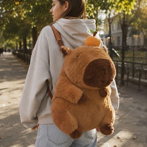 May include: A brown plush backpack shaped like a capybara, with a large snout and small ears. The backpack has adjustable brown straps and a small orange fruit on top. The backpack is worn by a person wearing a gray hoodie and blue jeans.