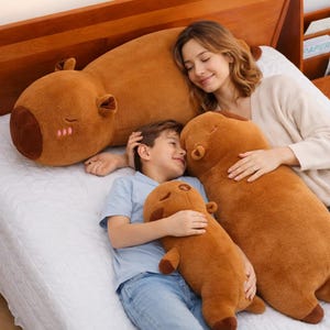 May include: A cozy scene featuring three brown capybara plush toys of varying sizes. The largest plush is positioned at the head of the bed, while a smaller one is held by a child. The toys have closed eyes and blush details.