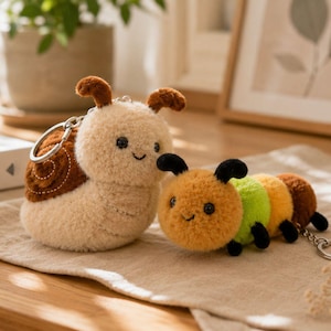 May include: Two plush keychains: a snail and a caterpillar. The snail is beige with a brown shell and brown antennae. The caterpillar is orange, green, yellow, and brown with black antennae and legs. Both have smiling faces.