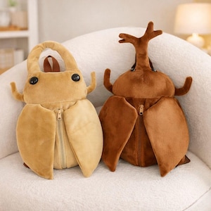 May include: Two plush beetle-shaped backpacks in tan and brown. Each has a zippered compartment, large black eyes, and antennae. The tan backpack has a handle, while the brown one has a large horn-like structure. They are sitting on a white, textured surface.