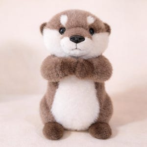 May include: A plush otter toy with a brown and white color scheme. The stuffed animal has a fluffy texture, with its arms crossed in front. The otter's face features a black nose and dark eyes, and it is sitting upright.