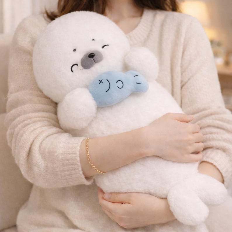 May include: A plush white seal toy with a light blue fish-shaped accessory. The seal has a smiling face with black eyes and a gray nose. The toy is soft and cuddly, perfect for children or as a decorative item.