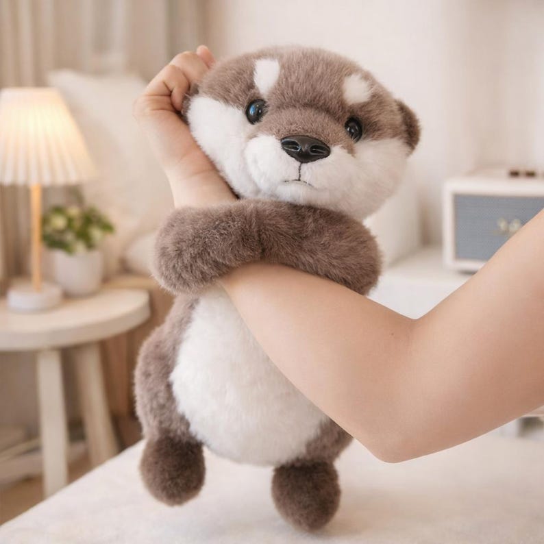 May include: A plush otter toy with a brown and white color scheme. The stuffed animal has a soft, furry texture and a cute, friendly expression. The toy is being held by a person.