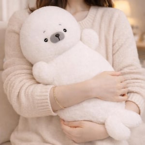May include: A plush white seal stuffed animal with a round head, small arms, and flipper-like feet. The seal has a gray nose and black eyes. The toy is soft and cuddly, perfect for children or as a decorative item.