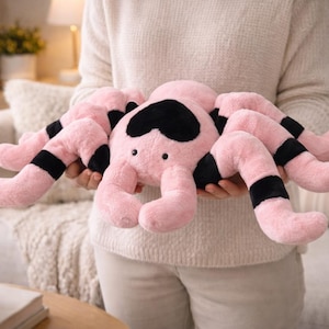 Puede incluir: Un peluche de araña rosa con detalles en negro. La araña tiene ocho patas rosas con bandas negras y una marca negra en forma de corazón en la cabeza. El peluche se sostiene frente a una persona, con un fondo neutro.
