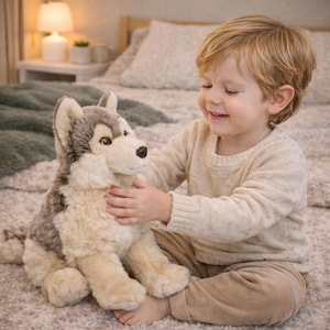 May include: A plush husky dog toy with gray and cream-colored fur. The toy has a black nose and is being held by a young child. The child is wearing a cream-colored sweater and tan pants. The background includes a bed and a lamp.