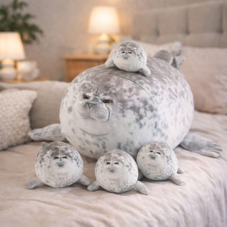 Pode incluir: Uma fam&iacute;lia de focas de pel&uacute;cia. Uma grande foca cinza e branca manchada com uma foca menor em cima e tr&ecirc;s focas menores na frente. Os bichos de pel&uacute;cia s&atilde;o macios e projetados para serem abra&ccedil;ados.