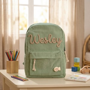 May include: A sage green corduroy backpack with the name "Wesley" embroidered in a light brown, rope-like font. The backpack has a front pocket, a small fish-shaped patch, and a tassel on the zipper. A wooden pencil holder and colored pencils are visible.
